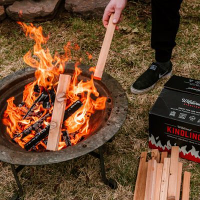 Person adding cedar kindling to a fire pit beside Wildwood Grilling kindling fire starters.